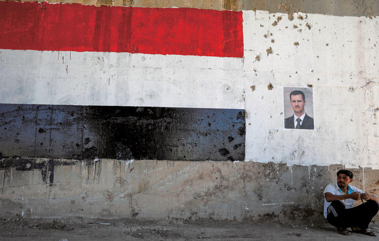 アサド大統領のポスターとその下に座り込む男性（昨年6月） OMAR SANADIKI-REUTERS