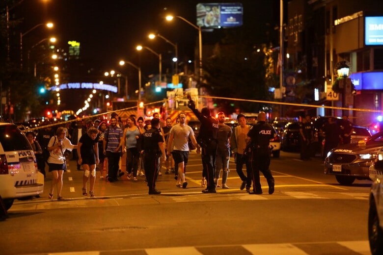 ７月２２日、カナダのトロントで、少女を含む１４人が銃撃され、うち１人が死亡した。現地警察のツイッターによると、容疑者は死亡した。負傷者の状態は分かっていない。規制線の張られた現地近くで遠巻きに様子を見る人たちと警備する警察官。トロントで撮影(２０１８年　ロイター／Chris Helgren)