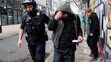 　米オレゴン州ポートランドの繁華街では、誰かが商店や流行最先端のレストランの前、ホテル付近、歩道、曲がり角、ベンチで身を屈めているのは見慣れた光景だ。手にしているのは、アルミホイル片や吸引パイプにかざしたライターだ。写真は、治療を受けるためのホットラインの電話番号が書かれたカードを薬物中毒者に渡す警察官（２０２４年　ロイター/Deborah Bloom）
