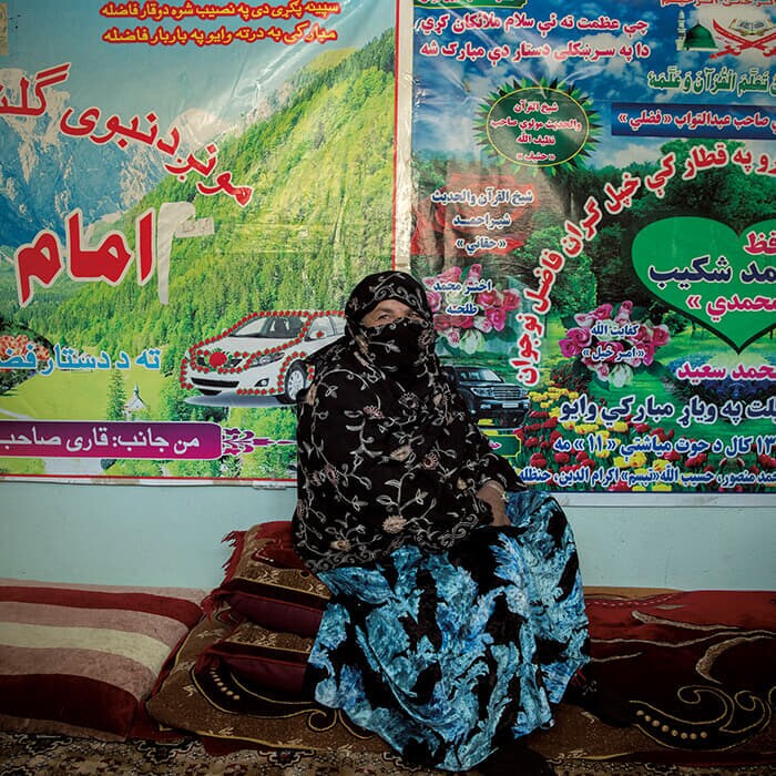 中部ワルダク州の自宅前でポーズを取る50歳のサイラ。壁に貼ってあるのは、息子たちがパキスタンのマドラサ(イスラム神学校)で入手したポスター。ワルダク州は内戦の影響が大きかったが、サイラは「内戦が終わった今は平和で静かだ」と語る