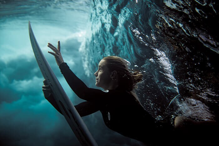 総合銀賞　「水泳」部門　『アンダーワールド』 »by Beatriz Ryder Da Costa　ソロモン諸島の海で波の下から浮かび上がるサーファー。ワニを警戒しながら夢中でシャッターを押し、想像もしなかった瞬間を切り取れたという