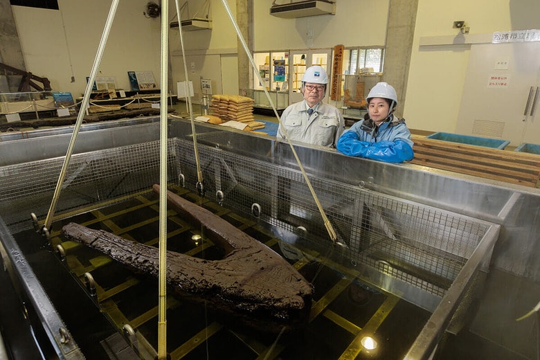 大型の含浸槽で保存処理が行われている元寇船の木製いかり（写真はクレーンで吊り上げた状態）。松浦市立埋蔵文化財センターでは伊藤幸司教授（左）の指導の下、職員の安木由美さん（同右）らが作業に当たる