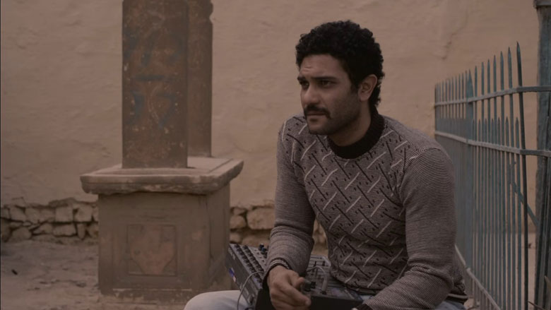 主人公が座る隣にある墓石。舞台になった場所は広大な墓地に人々が住む「死者の町」と呼ばれる場所＝映画『敷物と掛布』より