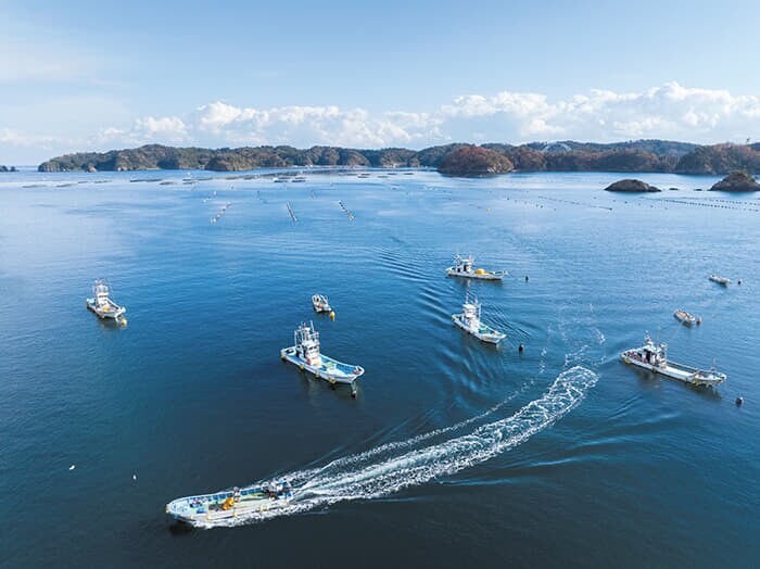 女川町尾浦（2024年）。ホタテやカキ、銀鮭の養殖が盛んな地区。作業を終えて帰港する船の姿と、陽光を受けたコバルトブルーの海が鮮烈に映る
